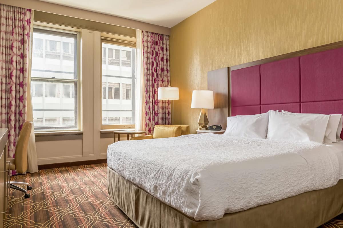 A hotel room with a large bed, a desk, a chair, a lamp, and two windows with curtains.