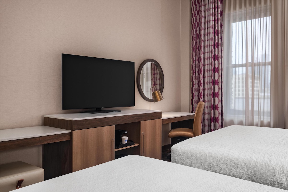 A hotel room with a flat-screen TV, a round mirror, a wooden desk, a chair, and two beds.