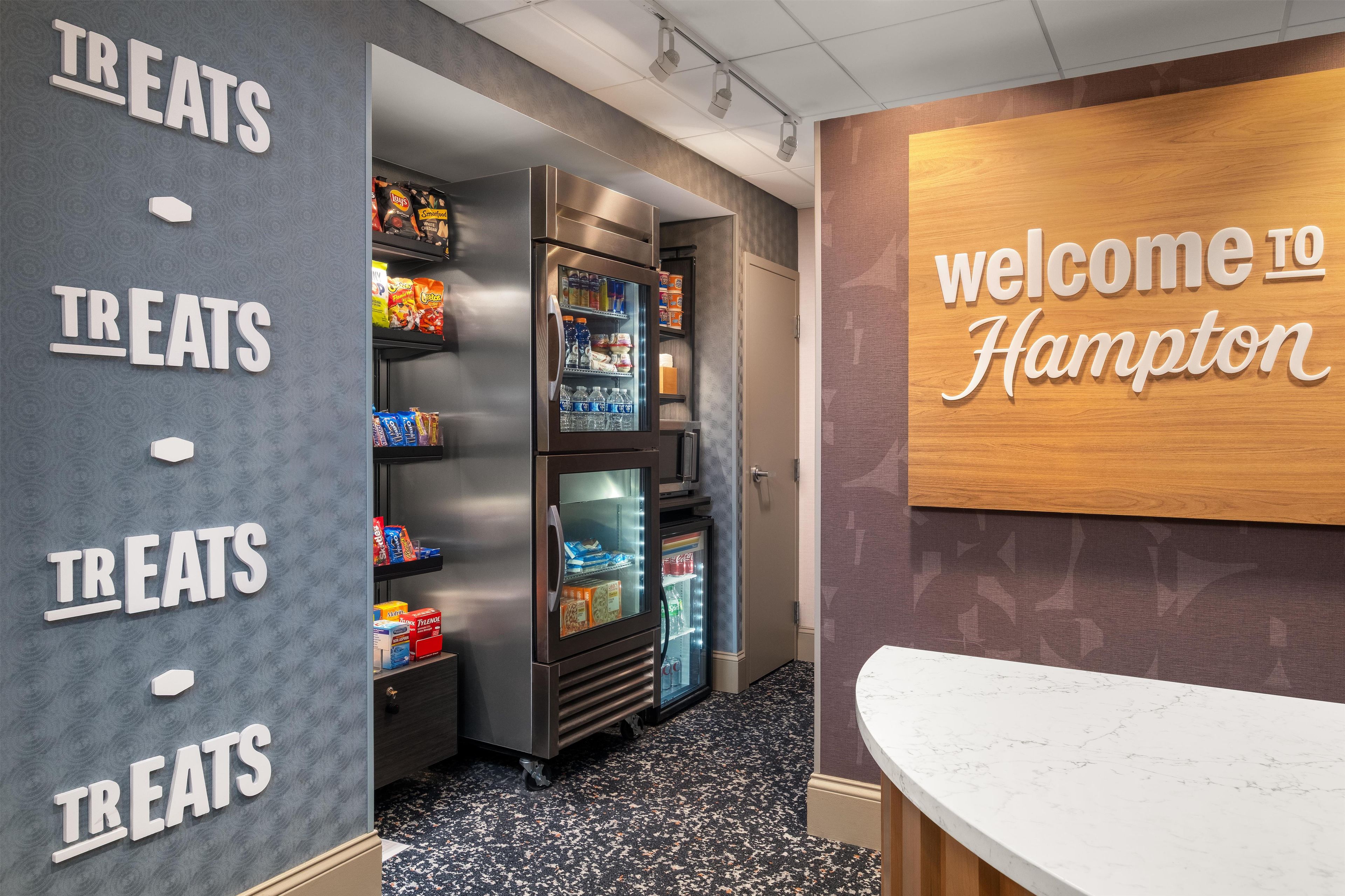 A hotel reception area with a wall sign reading 'welcome to Hampton' and a wall-mounted sign indicating a treats section.