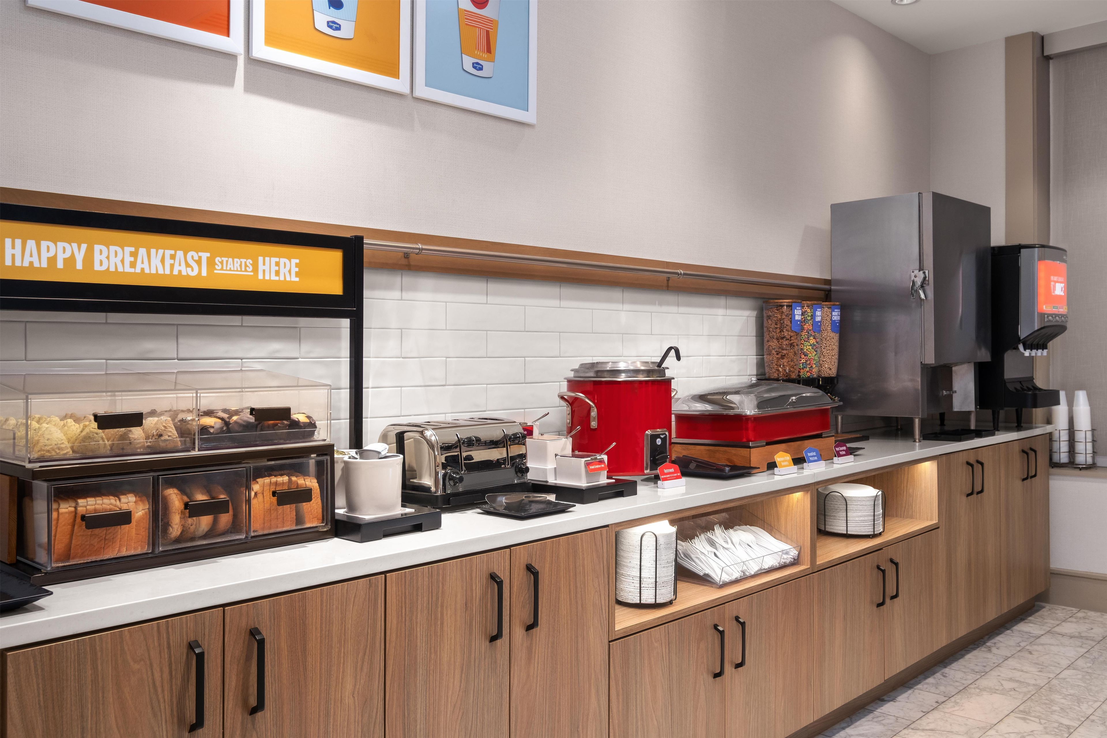 A breakfast buffet area with a sign reading 'HAPPY BREAKFAST STARTS HERE,' a toaster, a coffee machine, and various food items.