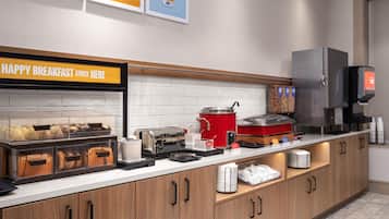 A breakfast buffet area with a sign reading 'HAPPY BREAKFAST STARTS HERE,' a toaster, a coffee machine, and various food items.