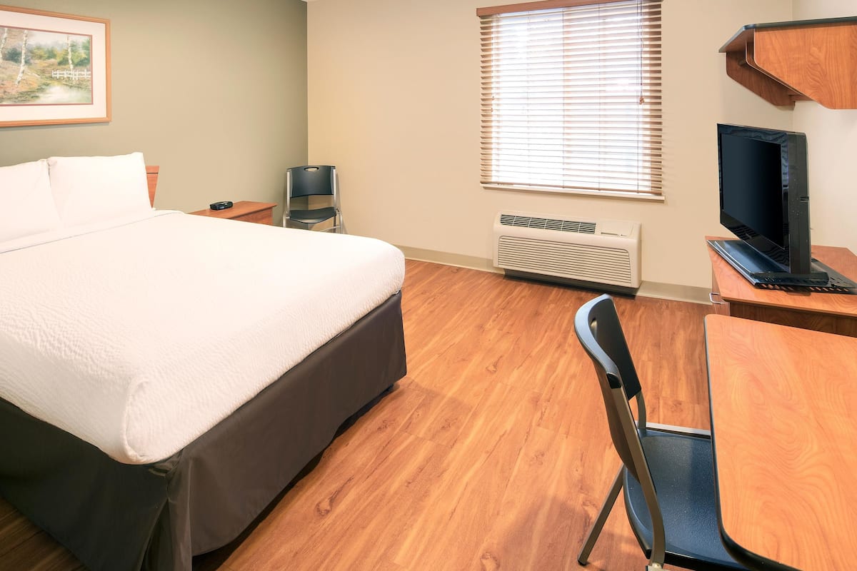Ein Hotelzimmer mit einem Bett, einem Schreibtisch mit Computer, einem Sessel, einem Fernseher und einem Fenster mit Jalousien.