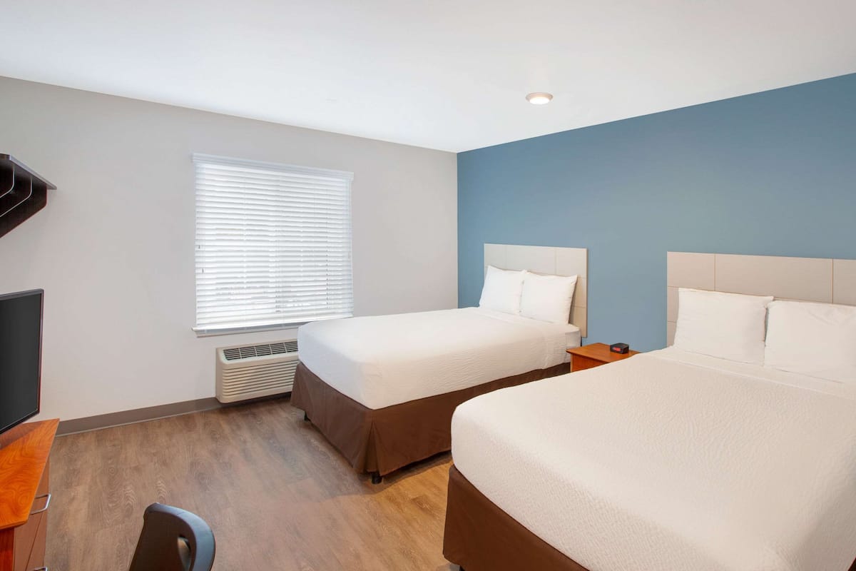 Ein Hotelzimmer mit zwei Betten, einem Schreibtisch, einem Sessel, einem Fernseher und einem Fenster mit Jalousien.