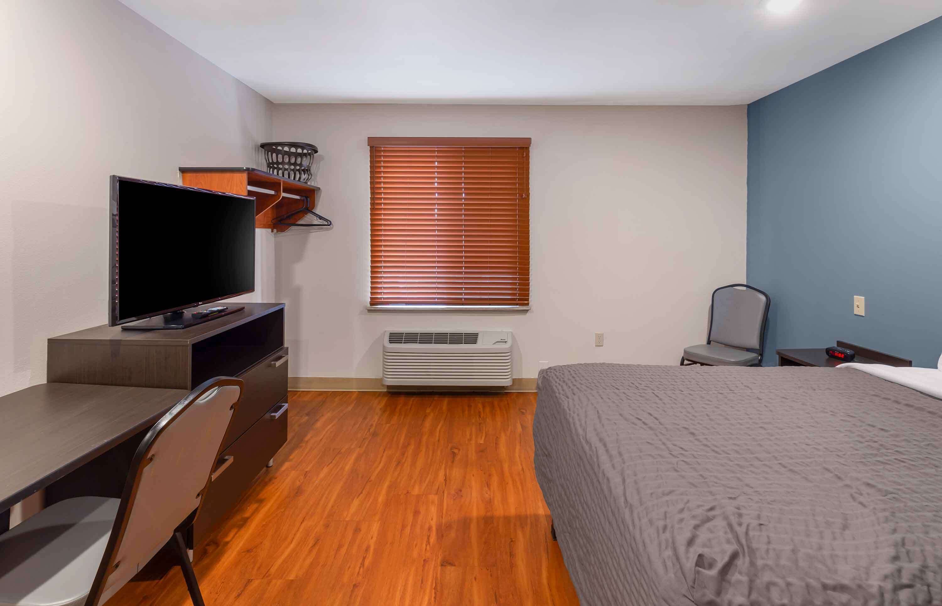 A hotel room with a bed, a desk with a TV, a chair, a window with blinds, and an air conditioner.