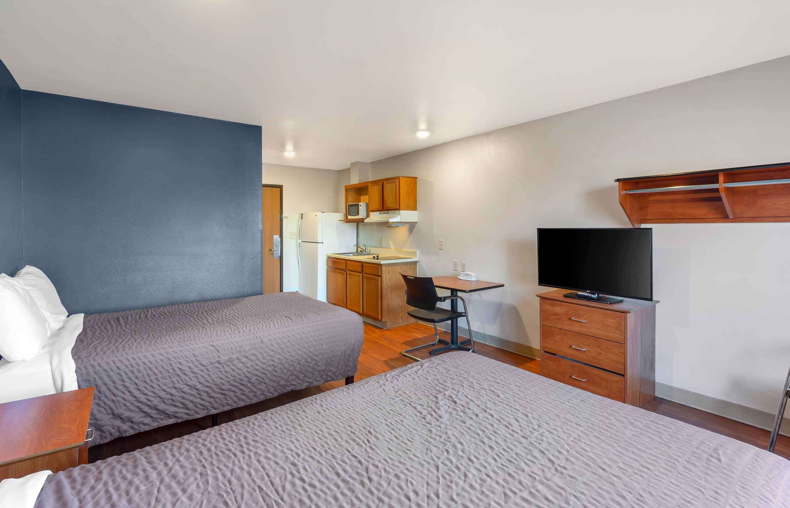 A hotel room with two beds, a desk with a chair, a TV, a microwave, and a small kitchenette.