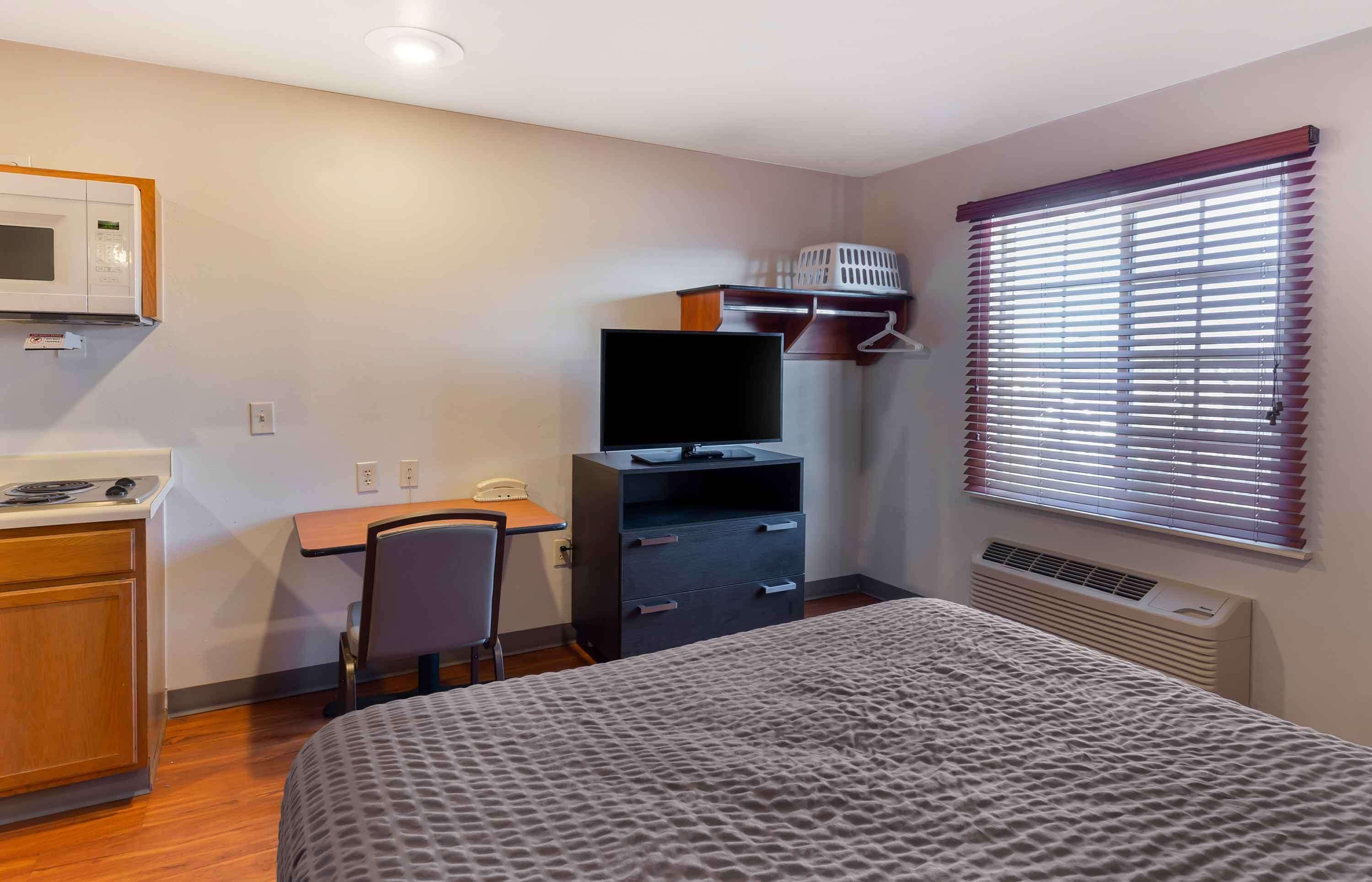 A hotel room with a bed, a desk with a computer, a microwave, a chair, and a window with blinds.
