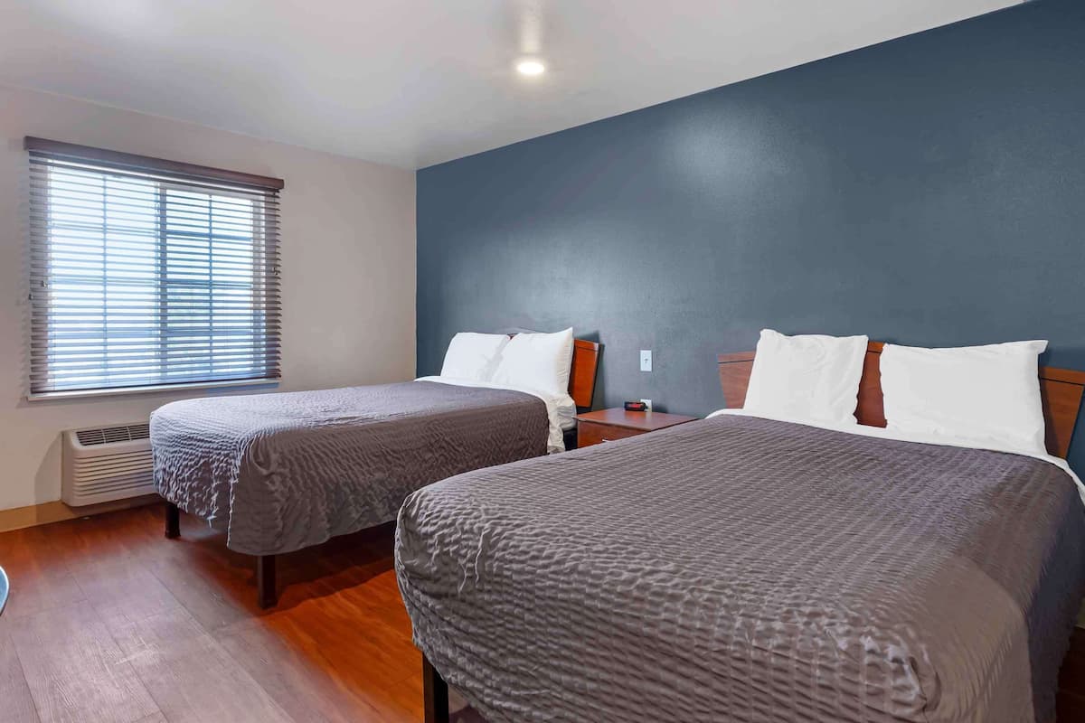 A hotel room with two beds, a window with blinds, a heater, and a blue accent wall.