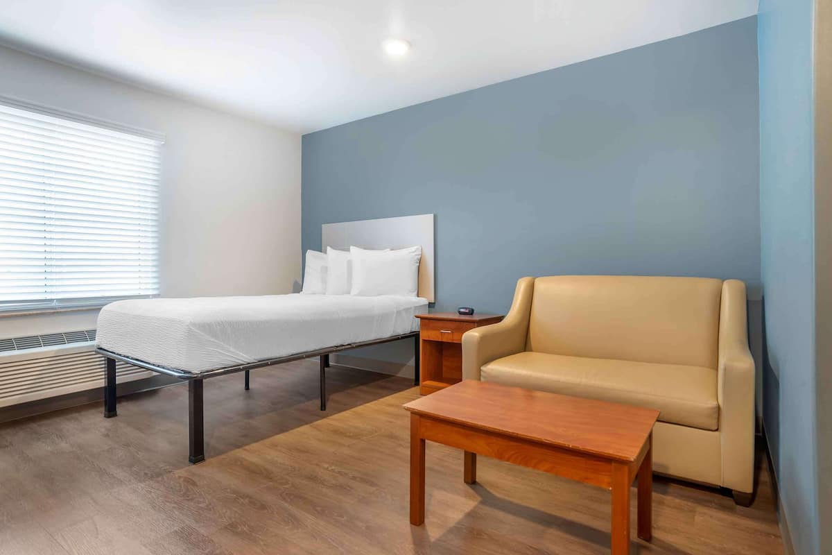 A hotel room with a bed, a chair, a small table, and a window with blinds.