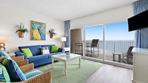 Una sala de estar moderna con un sofá azul, cojines verde y azul, una mesa de centro de vidrio y un gran ventanal con vista al océano.