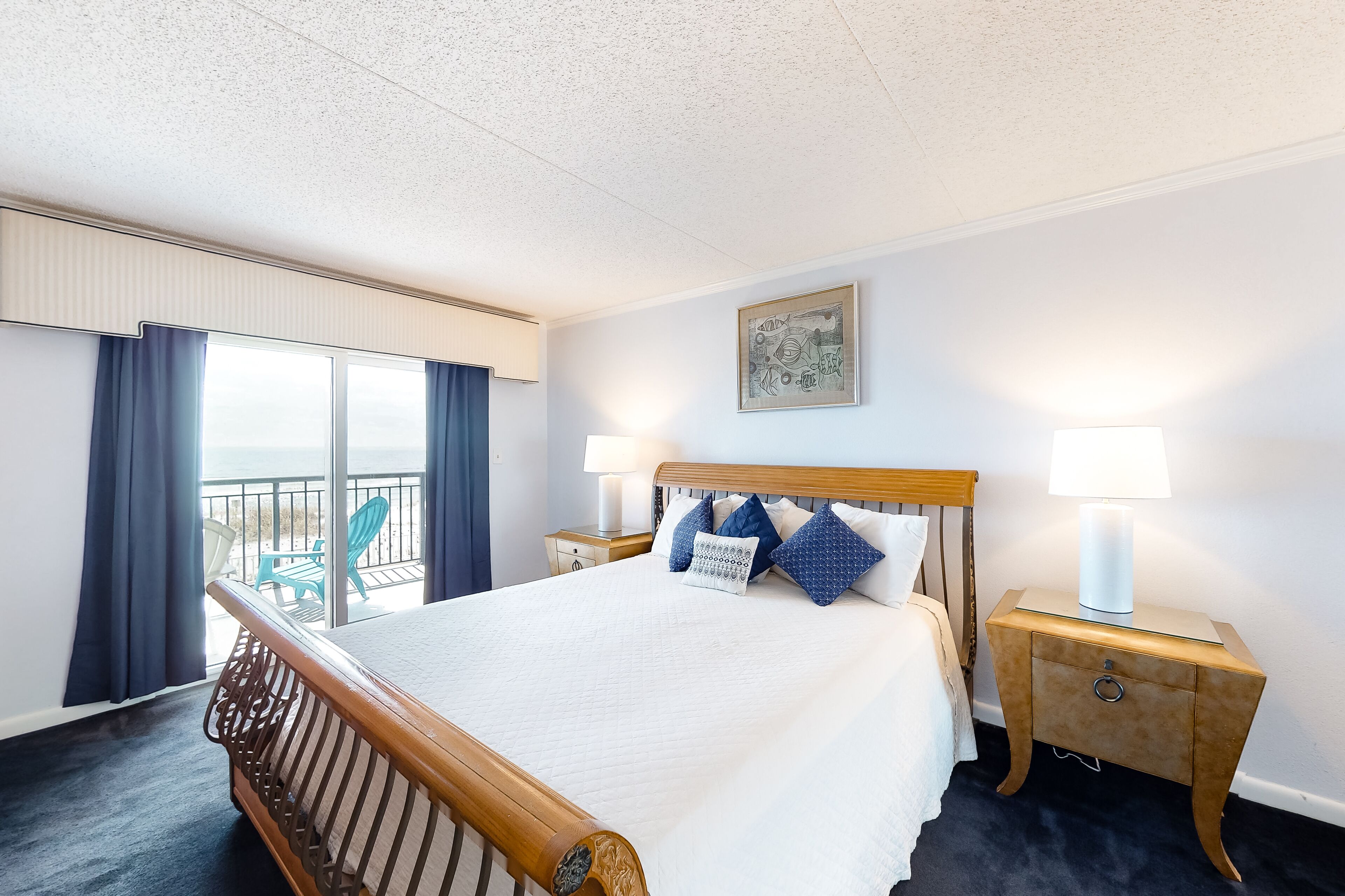 Habitación de hotel con una cama grande, dos mesitas de noche con lámparas, una ventana con cortinas, un balcón con sillas y un cuadro en la pared.
