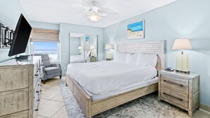 Une chambre à coucher comprenant un lit, un bureau, une chaise, une table de chevet, une lampe, un ventilateur de plafond, une fenêtre et un tableau.