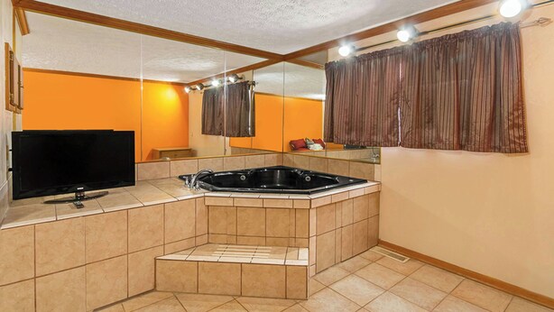Casa de banho com banheira, espelho, televisão e janela com cortinas.