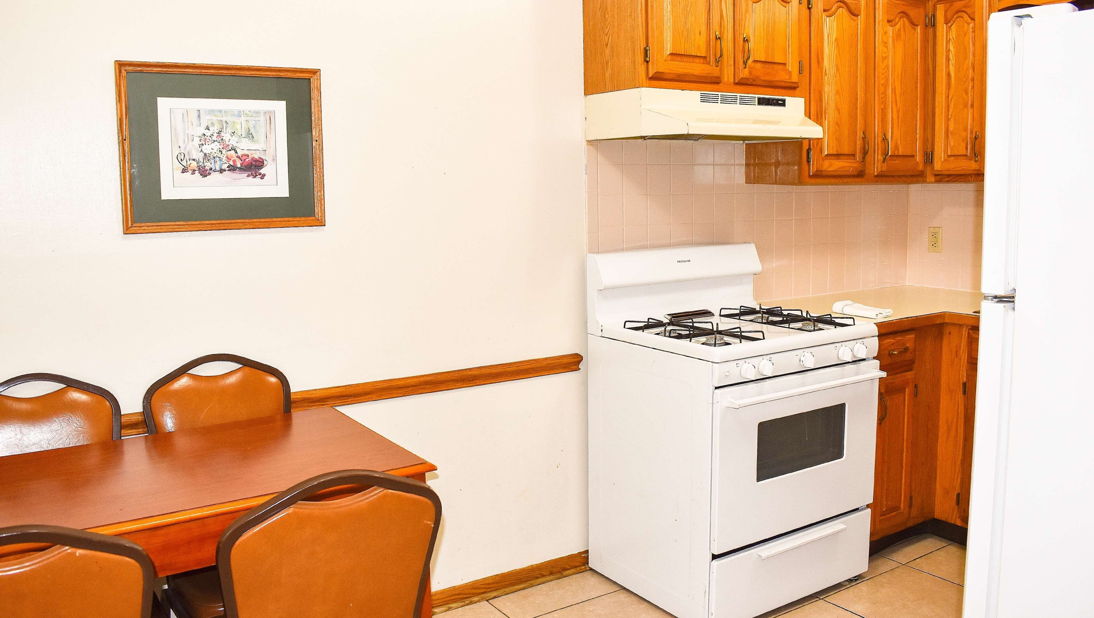 Cocina pequeña con estufa, horno y armarios de madera. Hay una mesa de comedor con sillas y un cuadro en la pared.