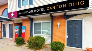 Um hotel com um letreiro luminoso indicando 'Magnuson Hotel Canton Ohio', fachada de tijolos, uma janela grande e duas máquinas de venda automática de Pepsi.