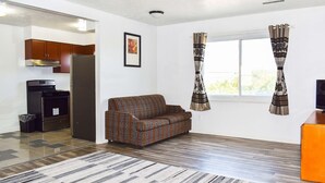 Uma sala de estar com sofá, mesa pequena, janela com cortinas e área de cozinha com armários e eletrodomésticos.