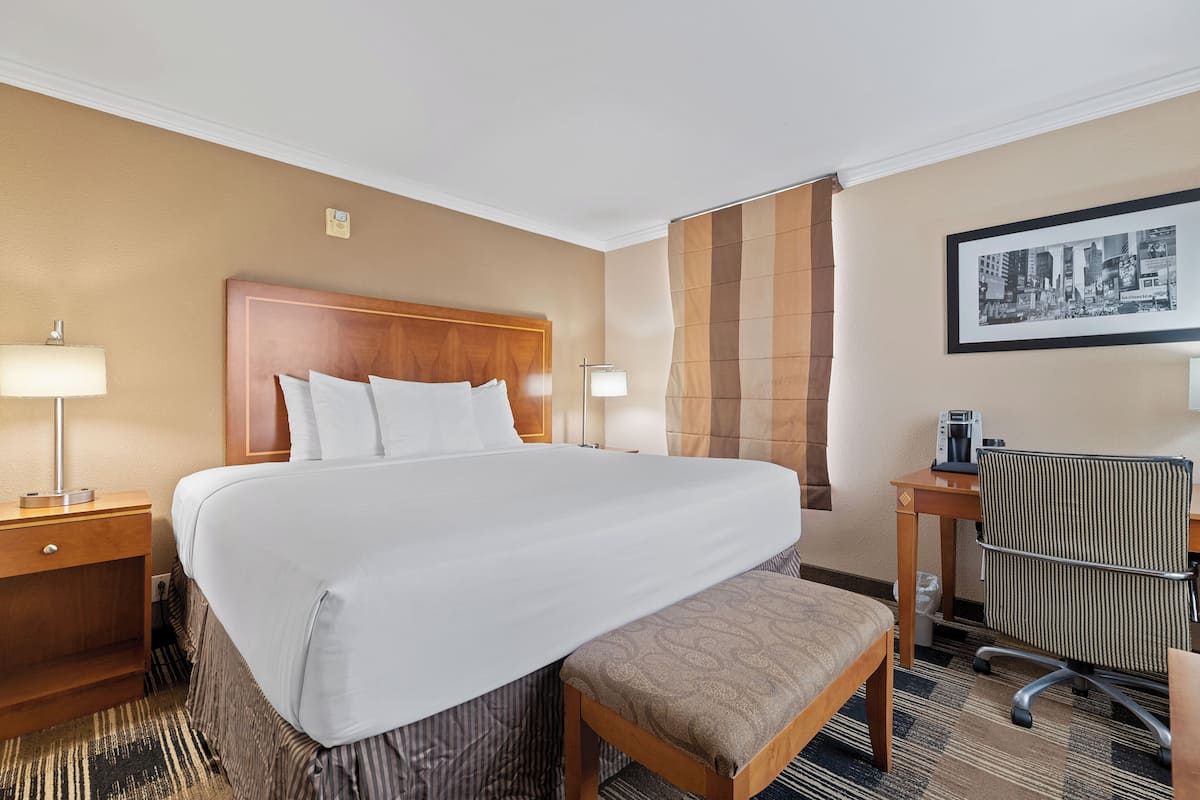 A hotel room with a large bed, bedside lamps, a desk with a chair, and a framed cityscape picture on the wall.