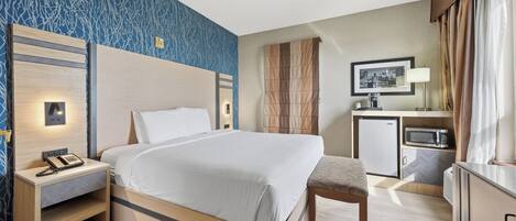 A hotel room with a large bed, bedside table with a phone, a desk with a microwave, and a chair.