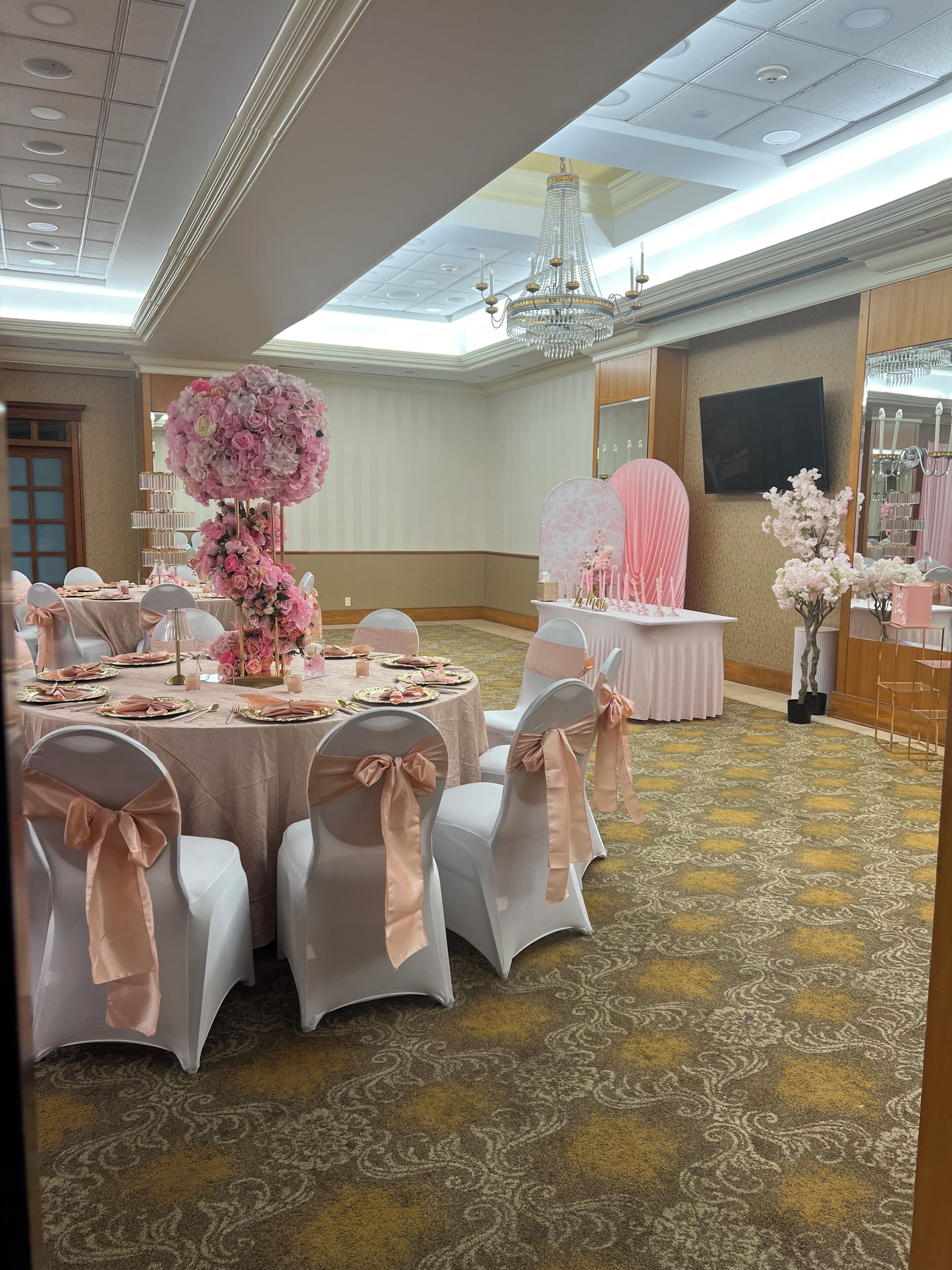 Un salón de banquetes con mesas redondas, sillas blancas adornadas con lazos y un gran centro de mesa floral.