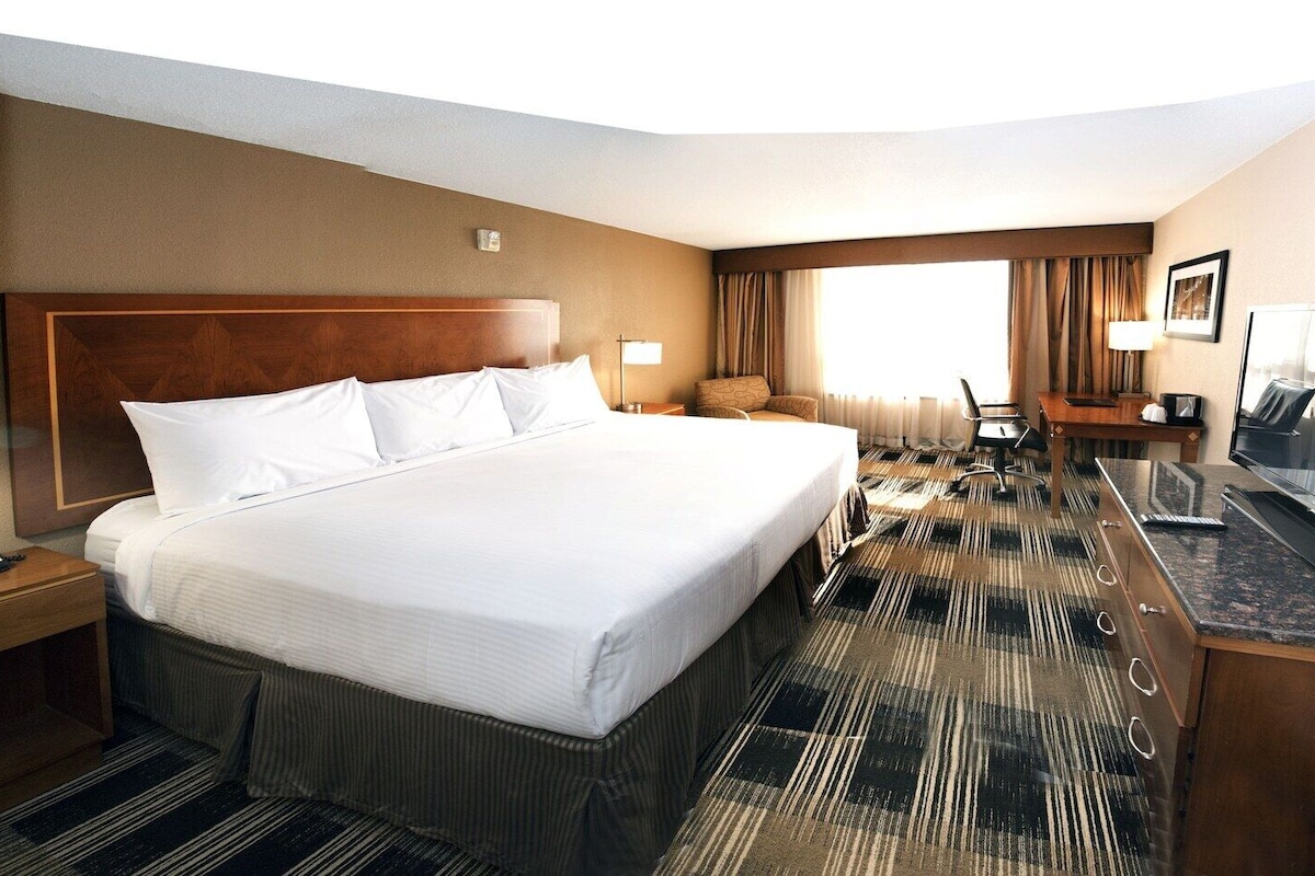 Ein Hotelzimmer mit einem großen Bett, einem Schreibtisch, einem Flachbildfernseher, einem Sessel, einer Lampe und einem Telefon.