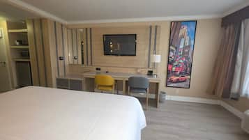 A hotel room with a large bed, a desk with two chairs, a television, and a cityscape painting on the wall.