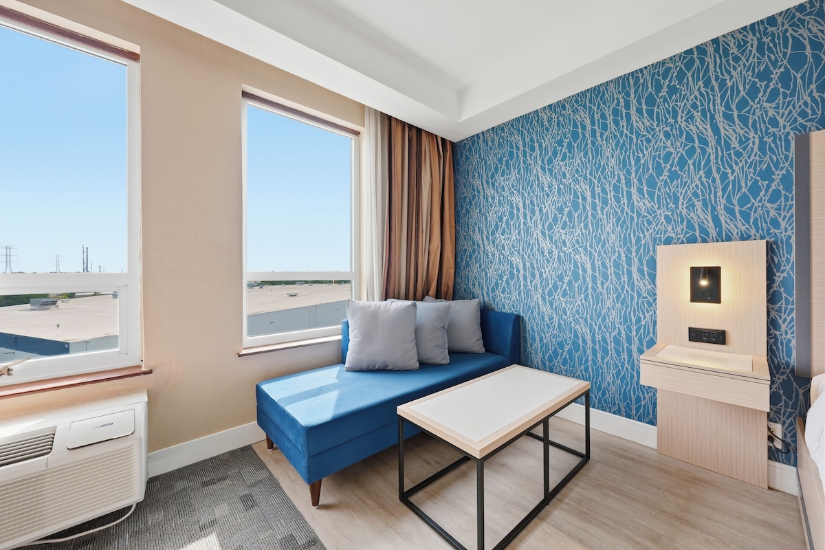 Ein Hotelzimmer mit einem blauen Sofa, einem kleinen Tisch, einem Fenster mit Blick auf das Flughafengelände und einer Wand mit blauem, abstrakten Muster.