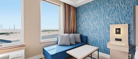 A hotel room with a blue sofa, a small table, a window with a view of the airport, and a wall with a blue abstract pattern.