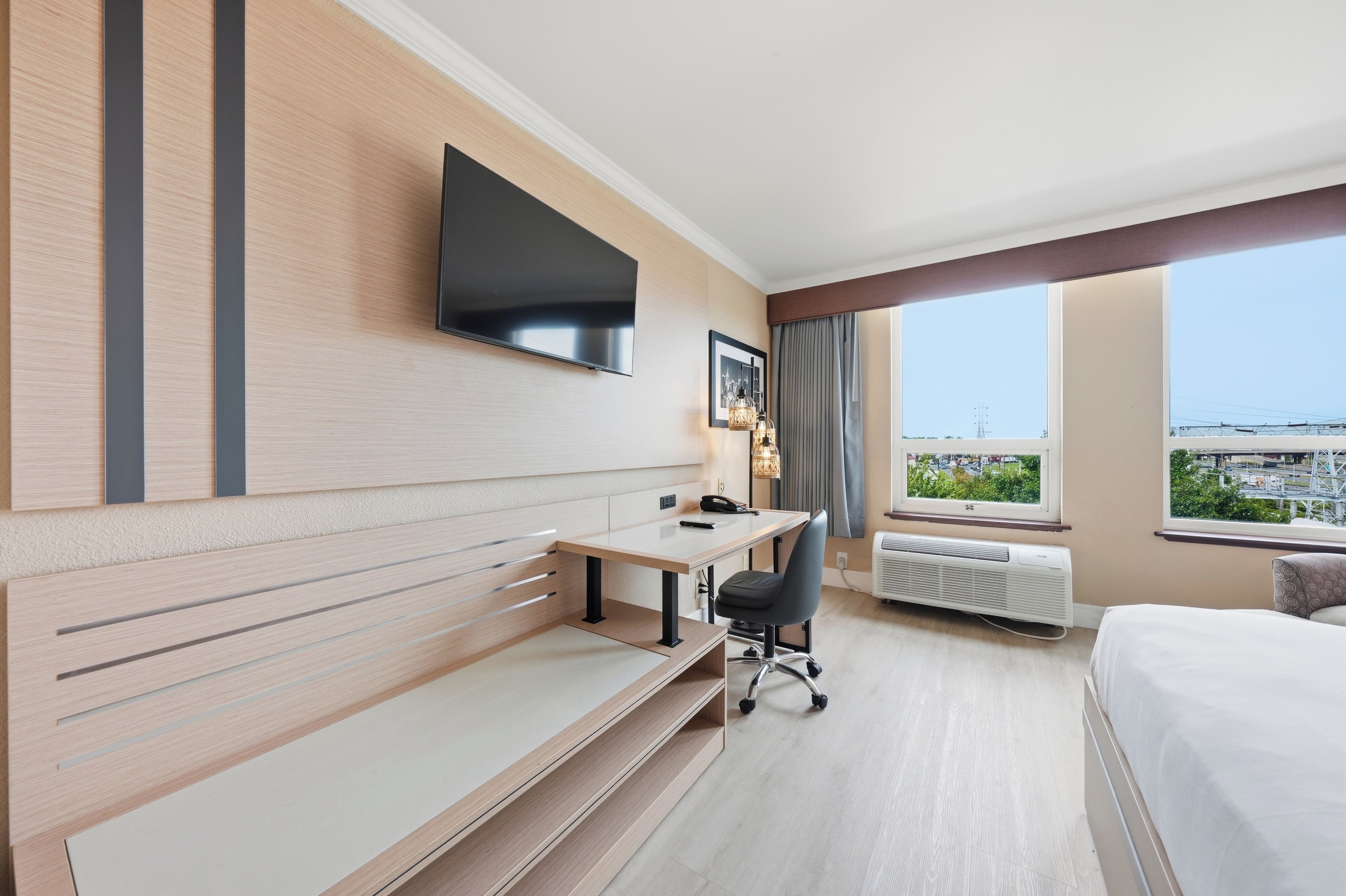 Quarto de hotel com cama, escrivaninha, cadeira, TV fixada na parede e uma grande janela com vista para a cidade.