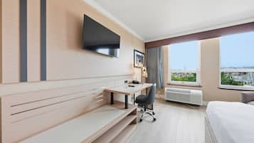 Quarto de hotel com cama, escrivaninha, cadeira, TV fixada na parede e uma grande janela com vista para a cidade.