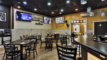 Een restaurantinterieur met meerdere tafels en stoelen, een aan de muur bevestigde televisie en een nooduitgangbord.