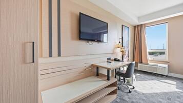 Habitación de hotel con televisor de pantalla plana montado en la pared, escritorio con silla, un banco, ventana con cortinas y vista a la ciudad.