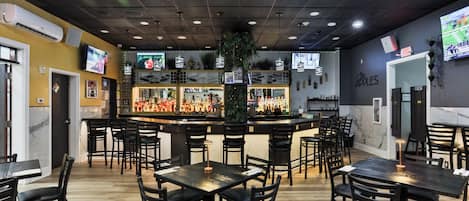 A restaurant interior with multiple tables and chairs, a bar area with bottles, and a television screen mounted on the wall.