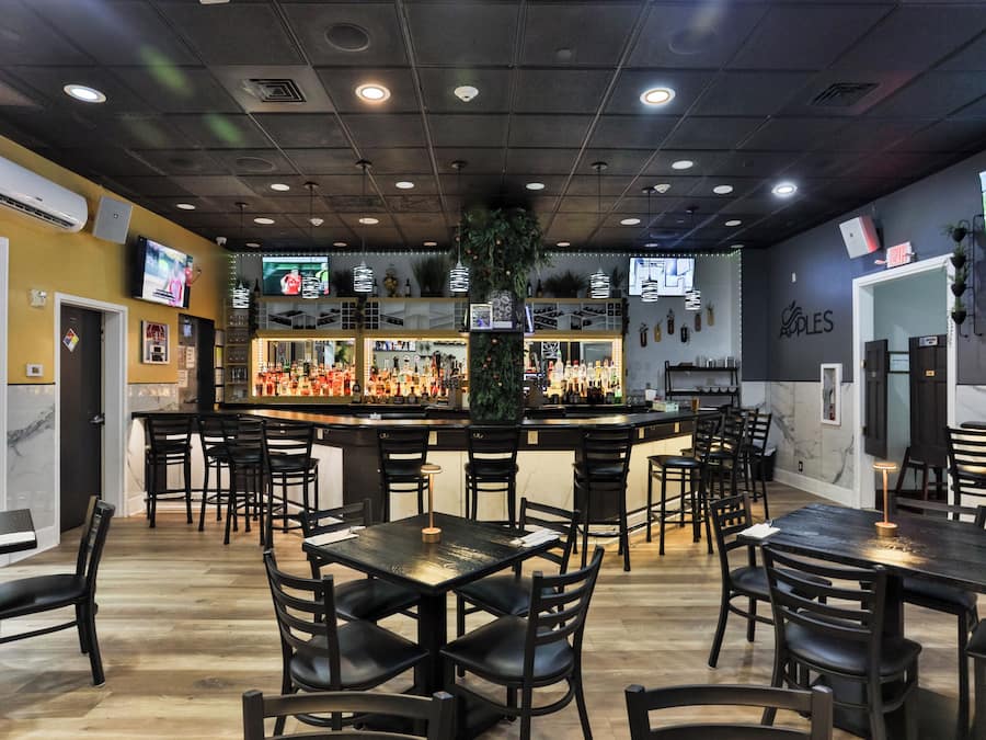 A restaurant interior with multiple tables and chairs, a bar area with bottles, and a television screen mounted on the wall.