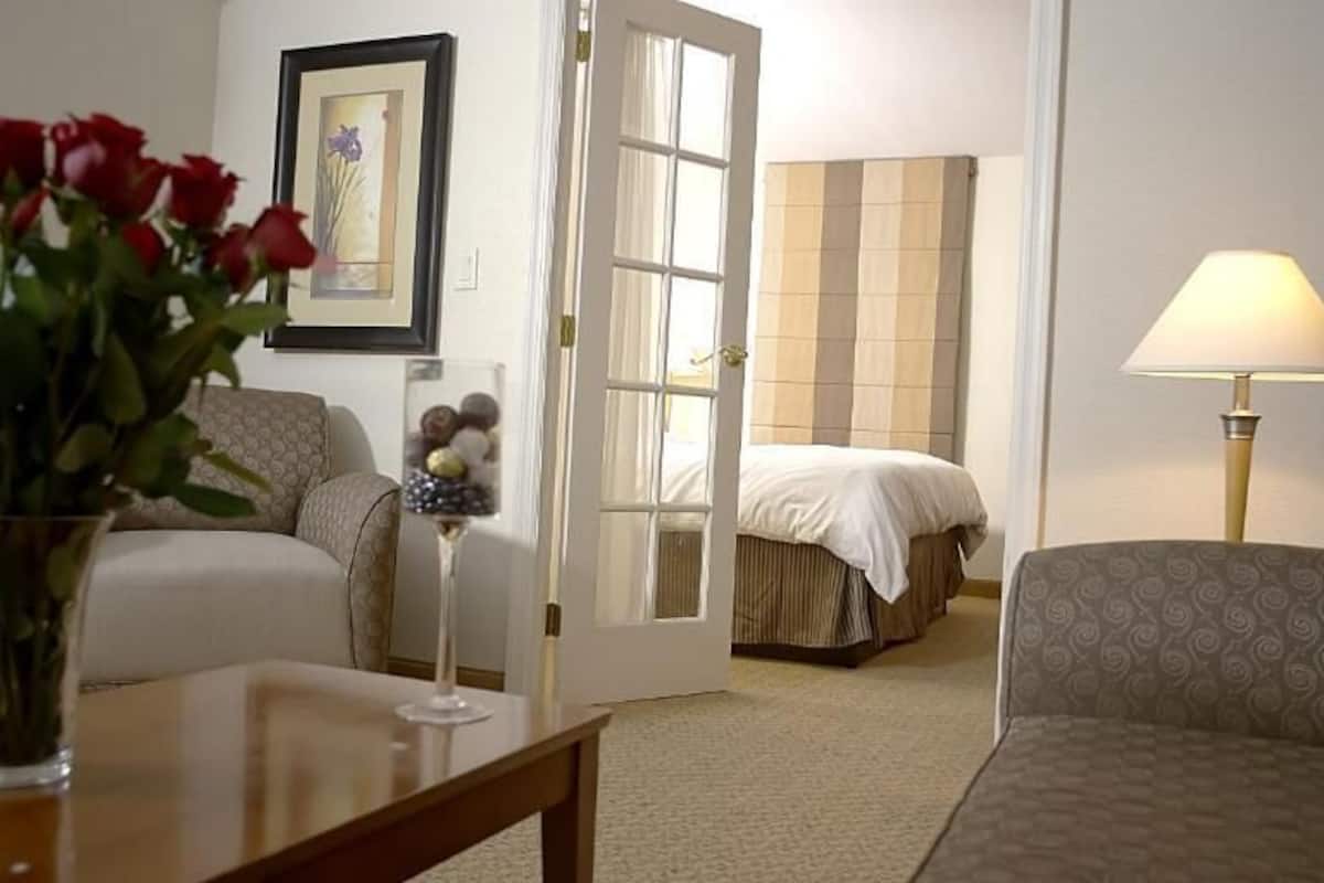 A hotel room with a bed, a sofa, a table with a vase of flowers, a lamp, and a door leading to another room.