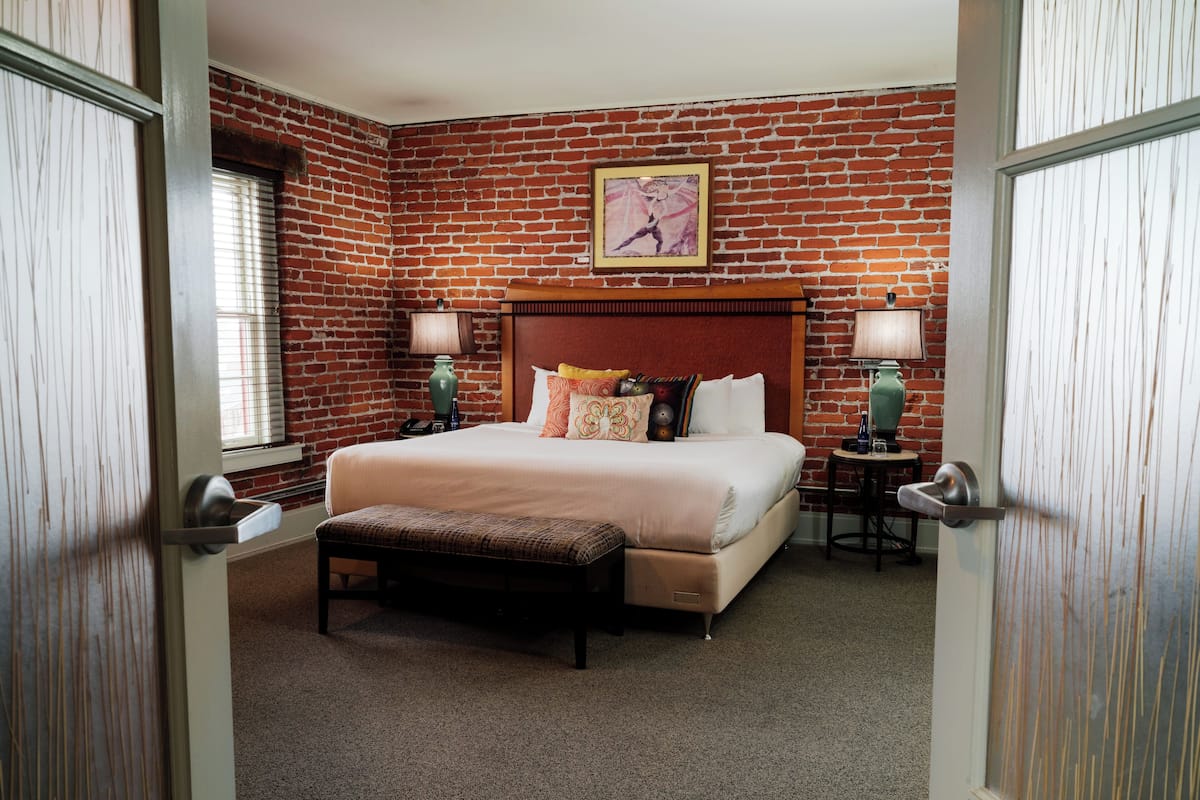 Habitación de hotel con pared de ladrillo, una cama grande con varias almohadas, dos mesitas de noche con lámparas y un banco al pie de la cama.