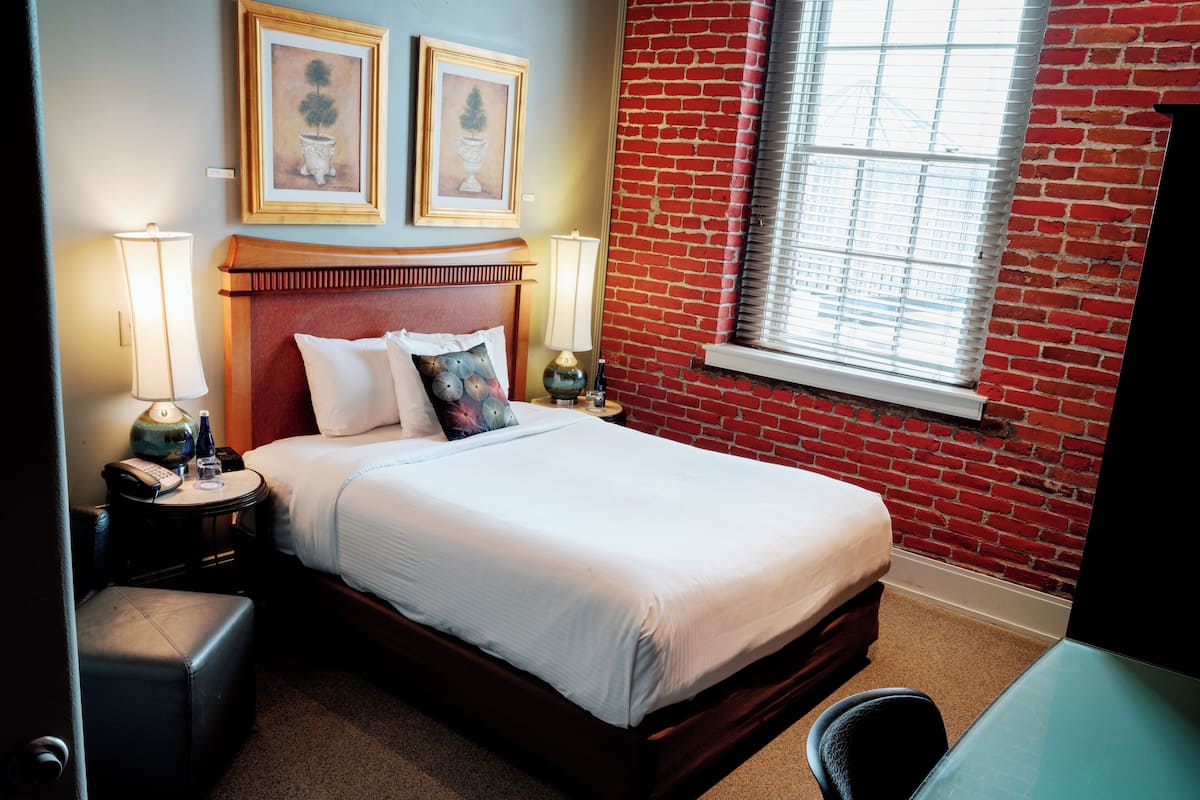 Una habitación de hotel con cama, mesita de noche, lámpara, silla y ventana con persianas.