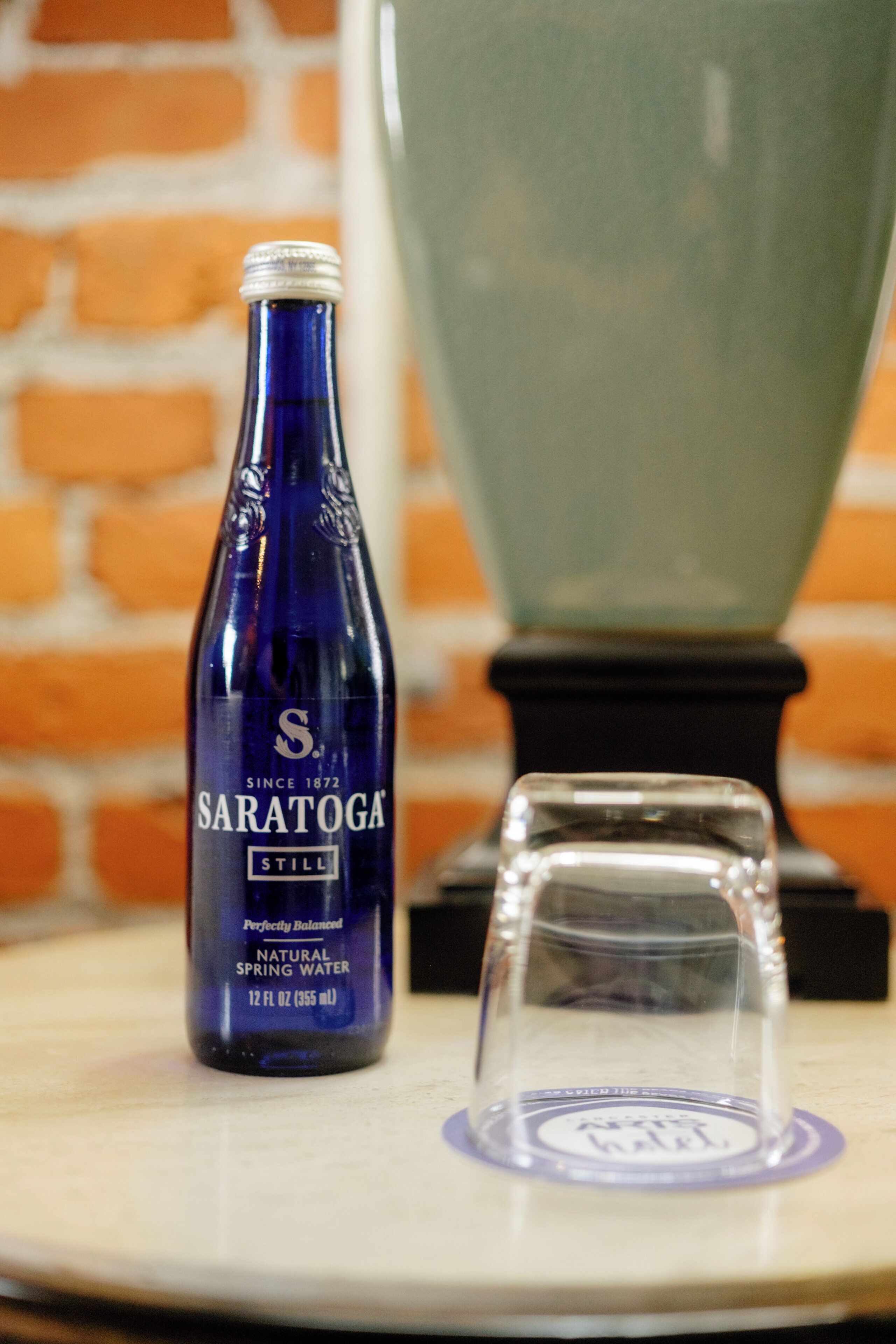 A blue bottle labeled 'Saratoga' and a clear glass on a table with a brick wall in the background.