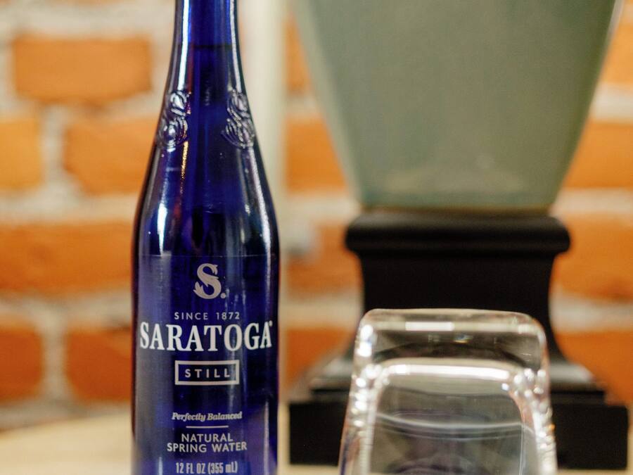 A blue bottle labeled 'Saratoga' and a clear glass on a table with a brick wall in the background.