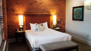 A hotel room with a brick wall, a bed with white linens, two bedside lamps, a bench, and a framed artwork on the wall.