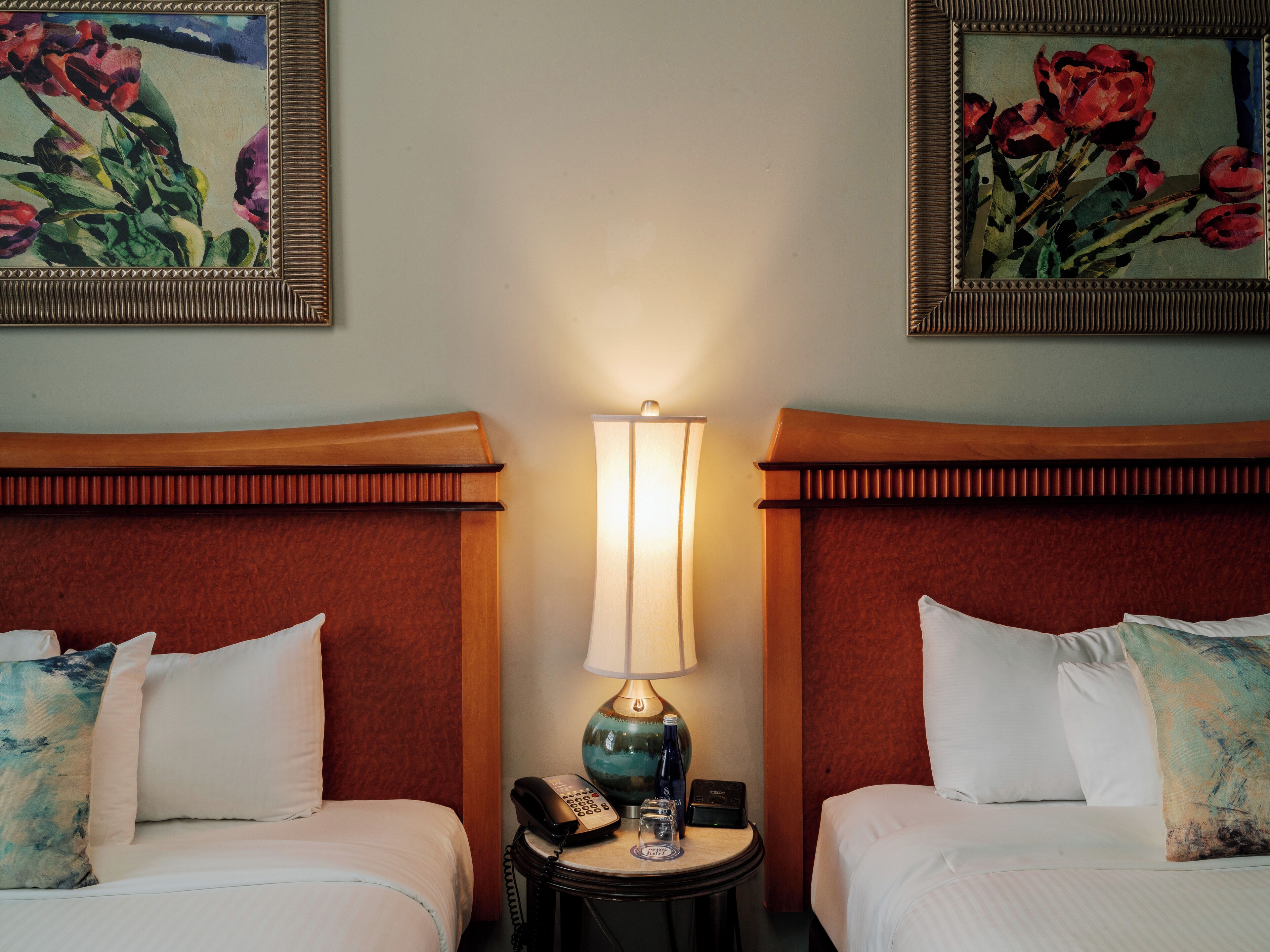 Quarto de hotel com duas camas, uma mesinha de cabeceira com abajur, telefone e uma pequena mesa com uma garrafa e um copo.