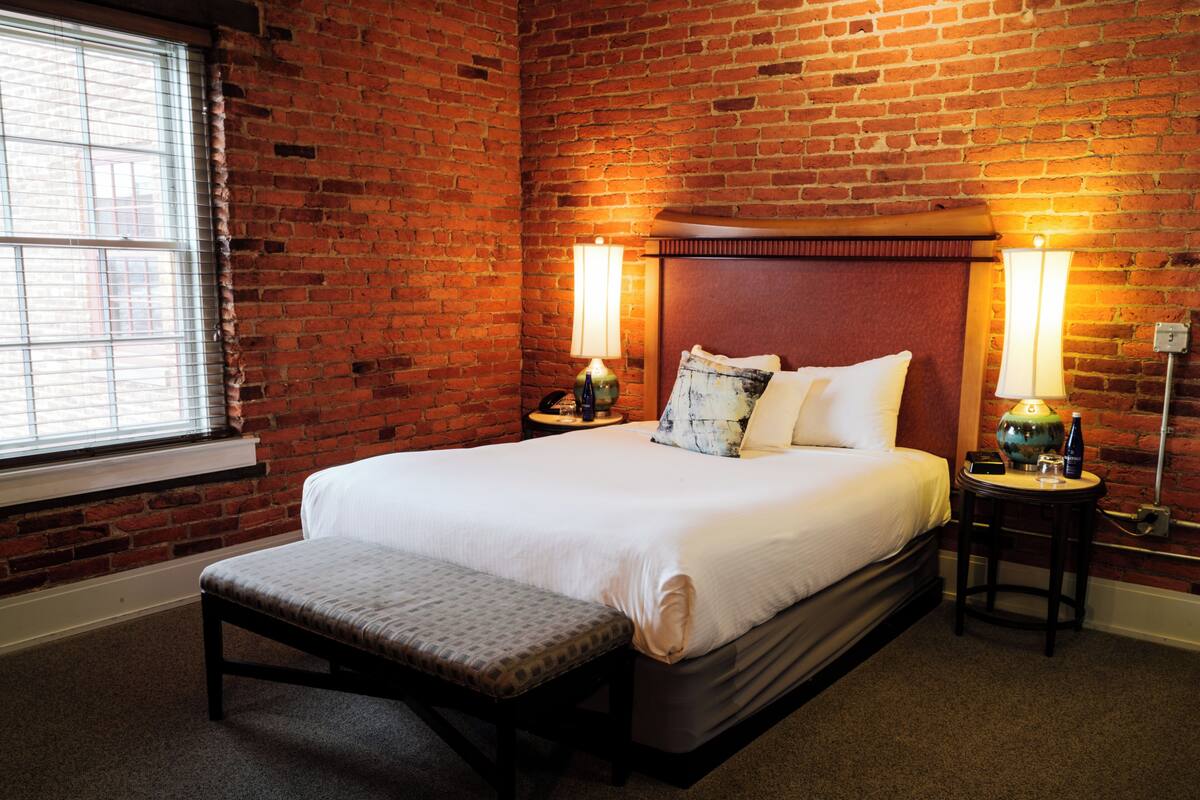 Una habitación de hotel con pared de ladrillo, una cama grande con ropa de cama blanca, dos mesitas de noche con lámparas y un banco al pie de la cama.