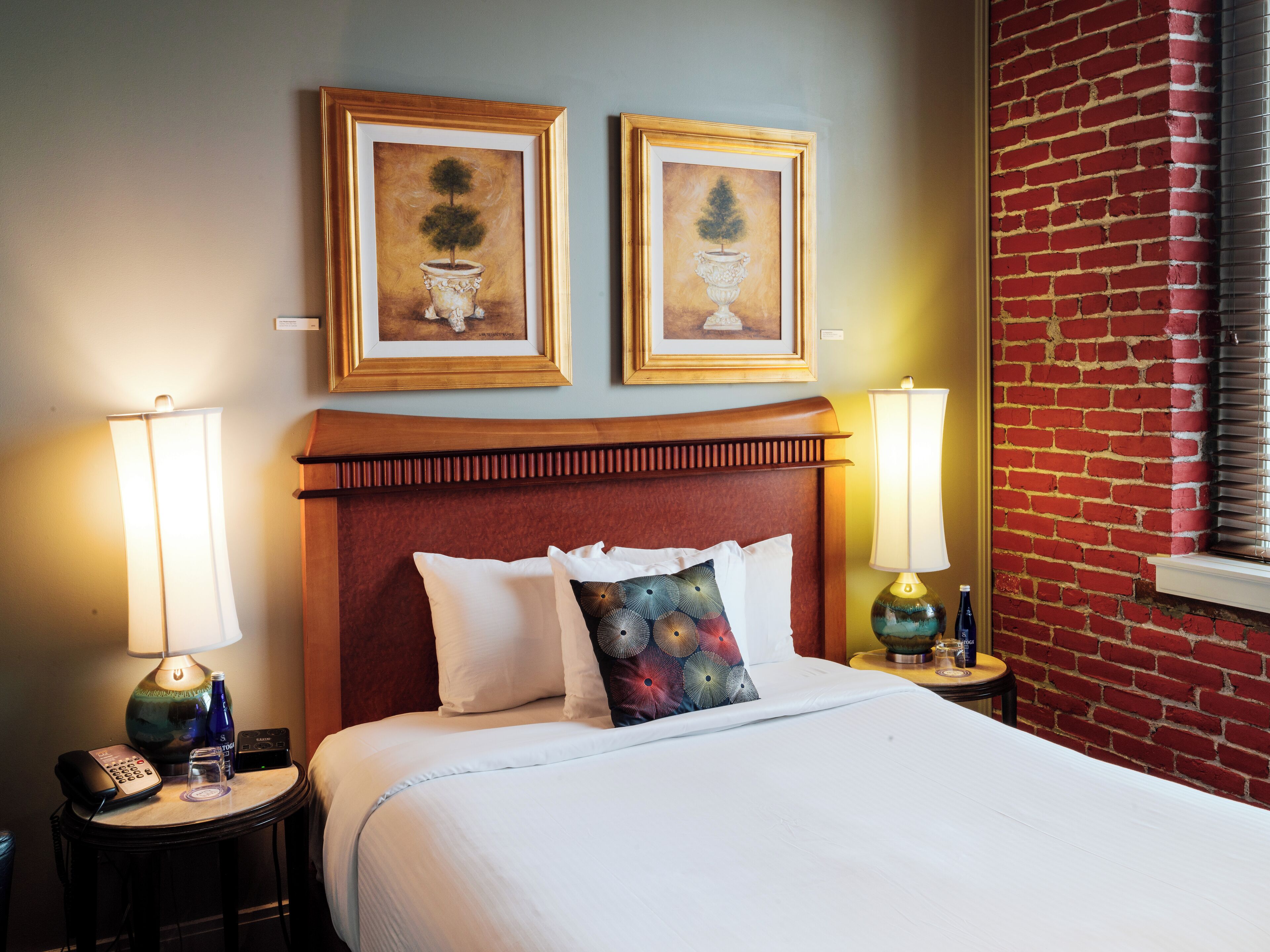 Quarto de hotel com uma cama, duas pinturas penduradas na parede, uma parede de tijolos vermelhos e duas luminárias de cabeceira.