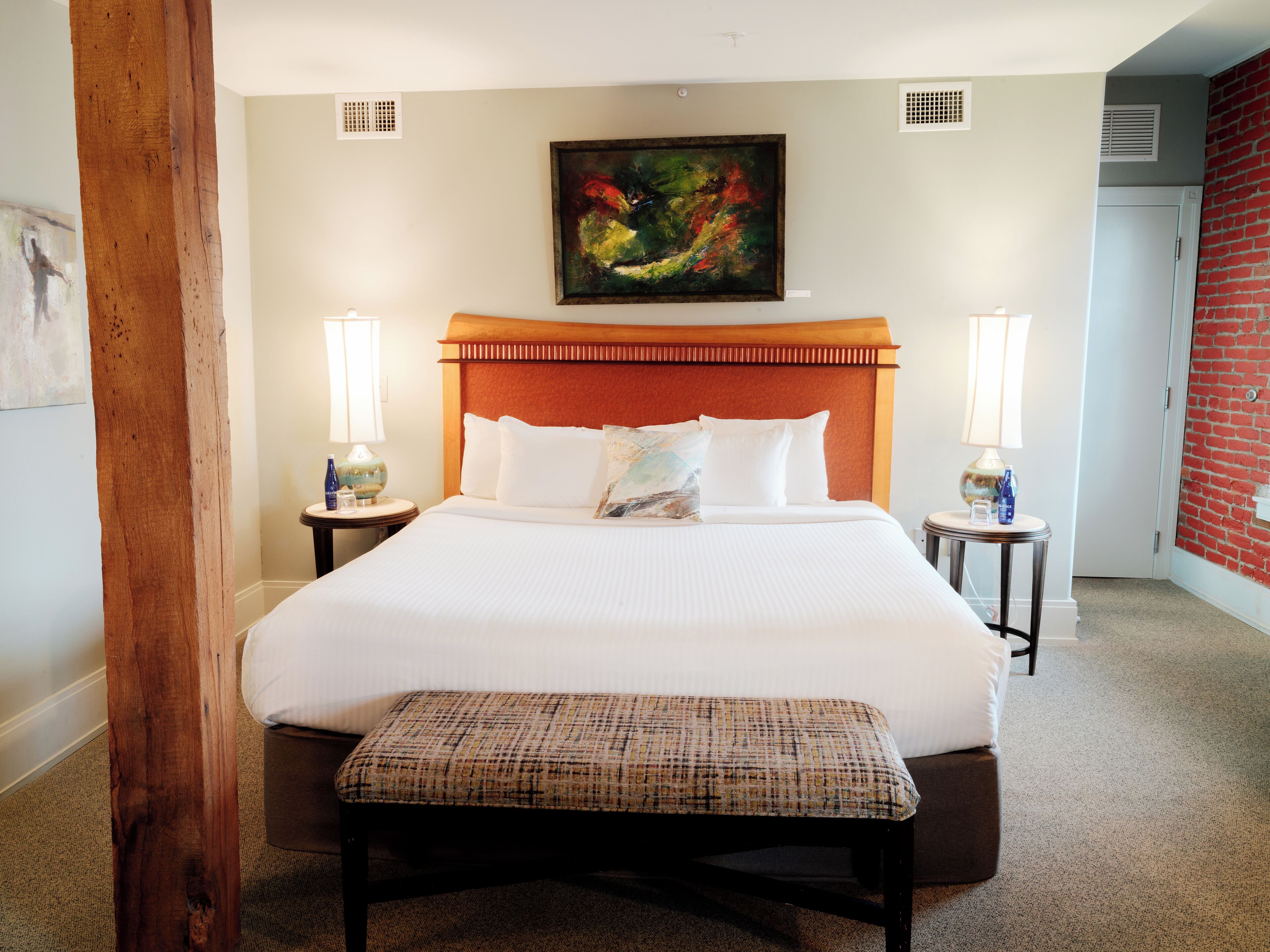 A hotel room with a large bed, two bedside tables with lamps, a bench, a painting on the wall, and a red brick wall.