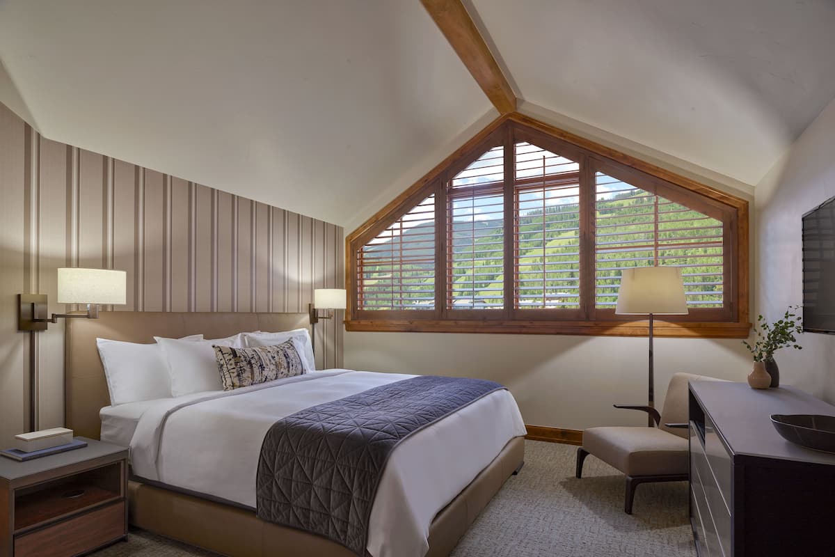 A bedroom with a large bed, two bedside tables, a desk, a chair, and a window with a view of greenery.