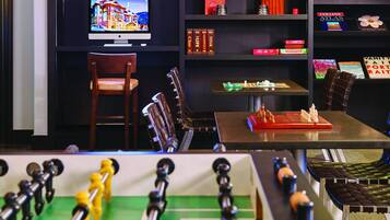 A room with a foosball table, a television displaying a scenic image, and shelves with books and board games.