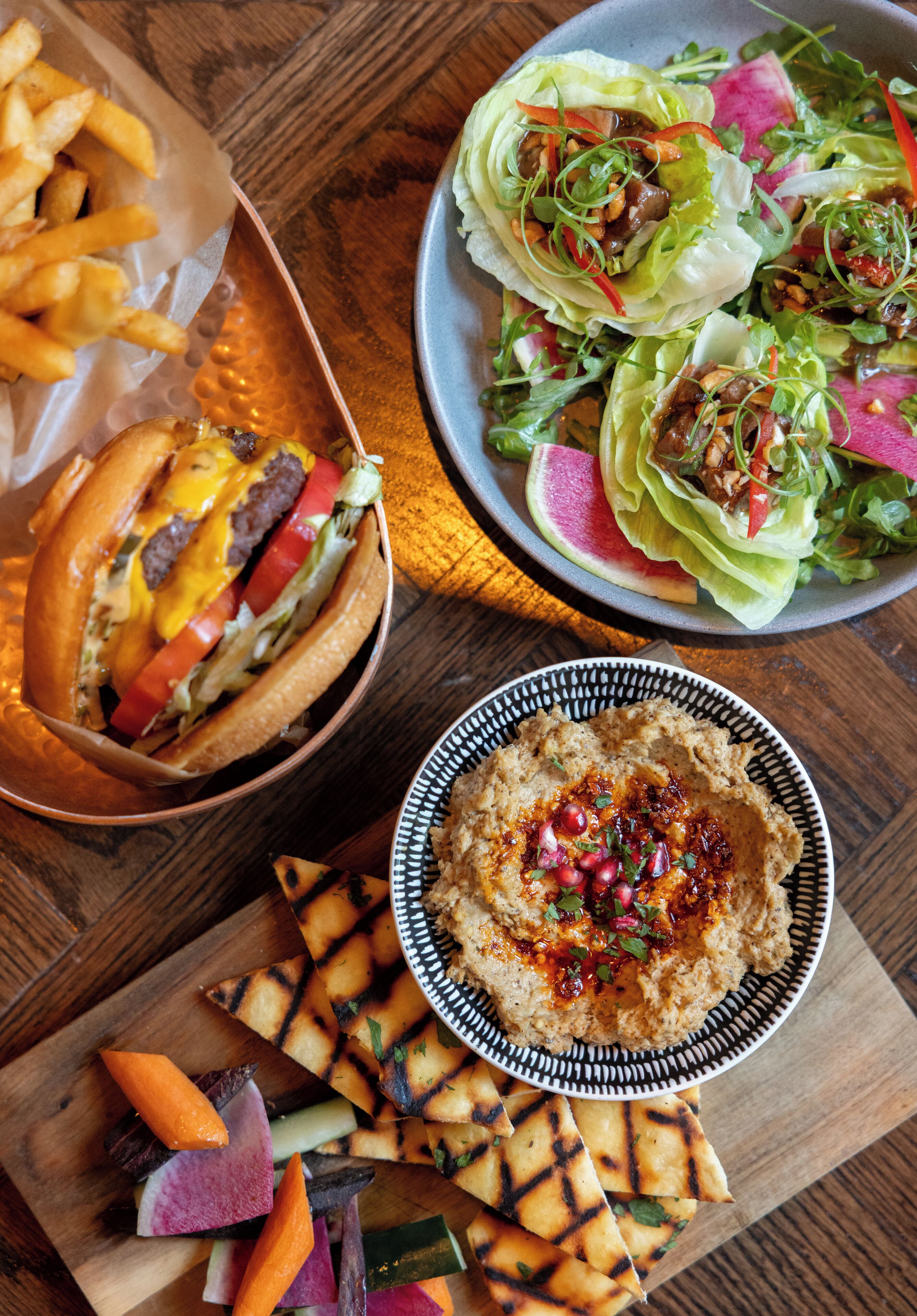 Una comida con hamburguesa, papas fritas, ensalada, hummus y verduras a la parrilla.