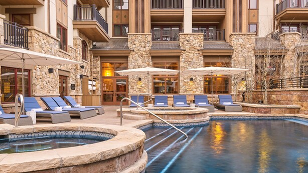 A pool area with lounge chairs and umbrellas, stone walls, and a multi-level building with balconies.