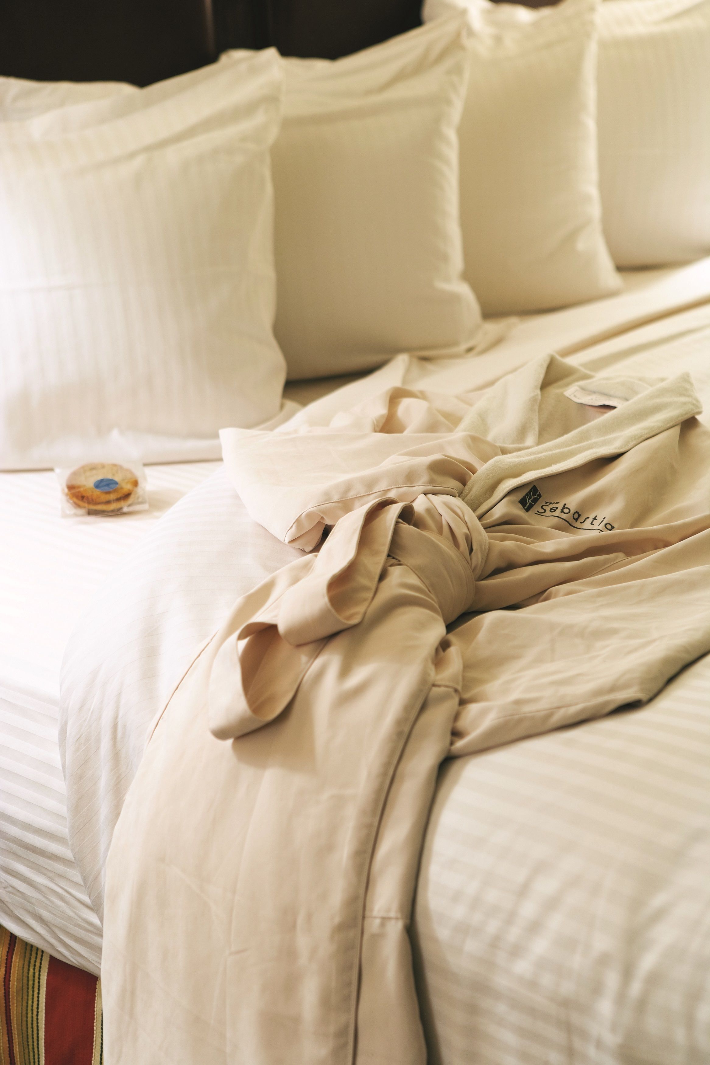A neatly made bed with a beige comforter and pillows, a small bag of snacks, and a piece of clothing with the word "Sebastian" on it.