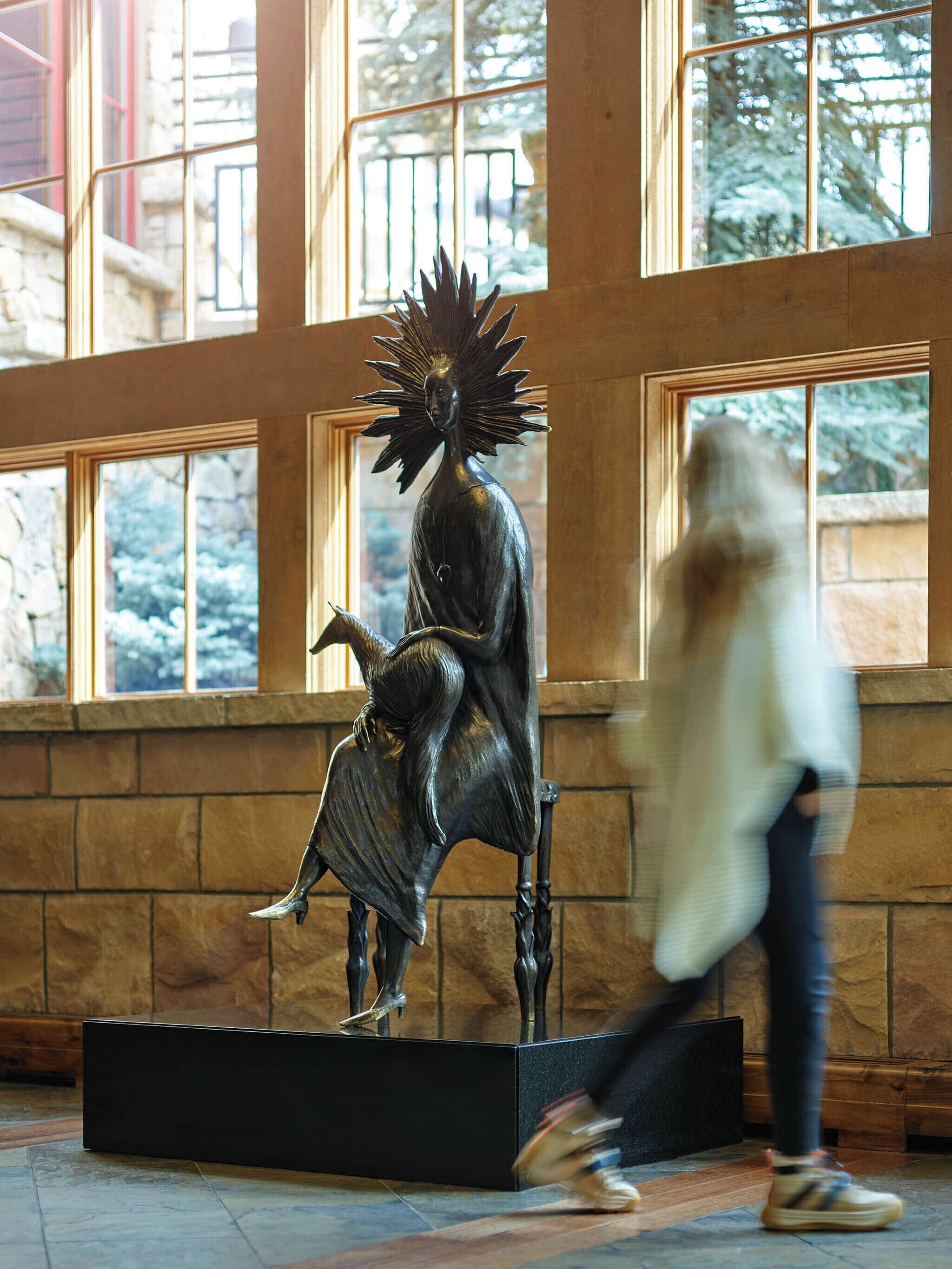 Une statue représentant une figure assise, coiffée d’un headdress orné de pointes, se trouve dans un hall d’entrée doté de grandes fenêtres et de murs en pierre.