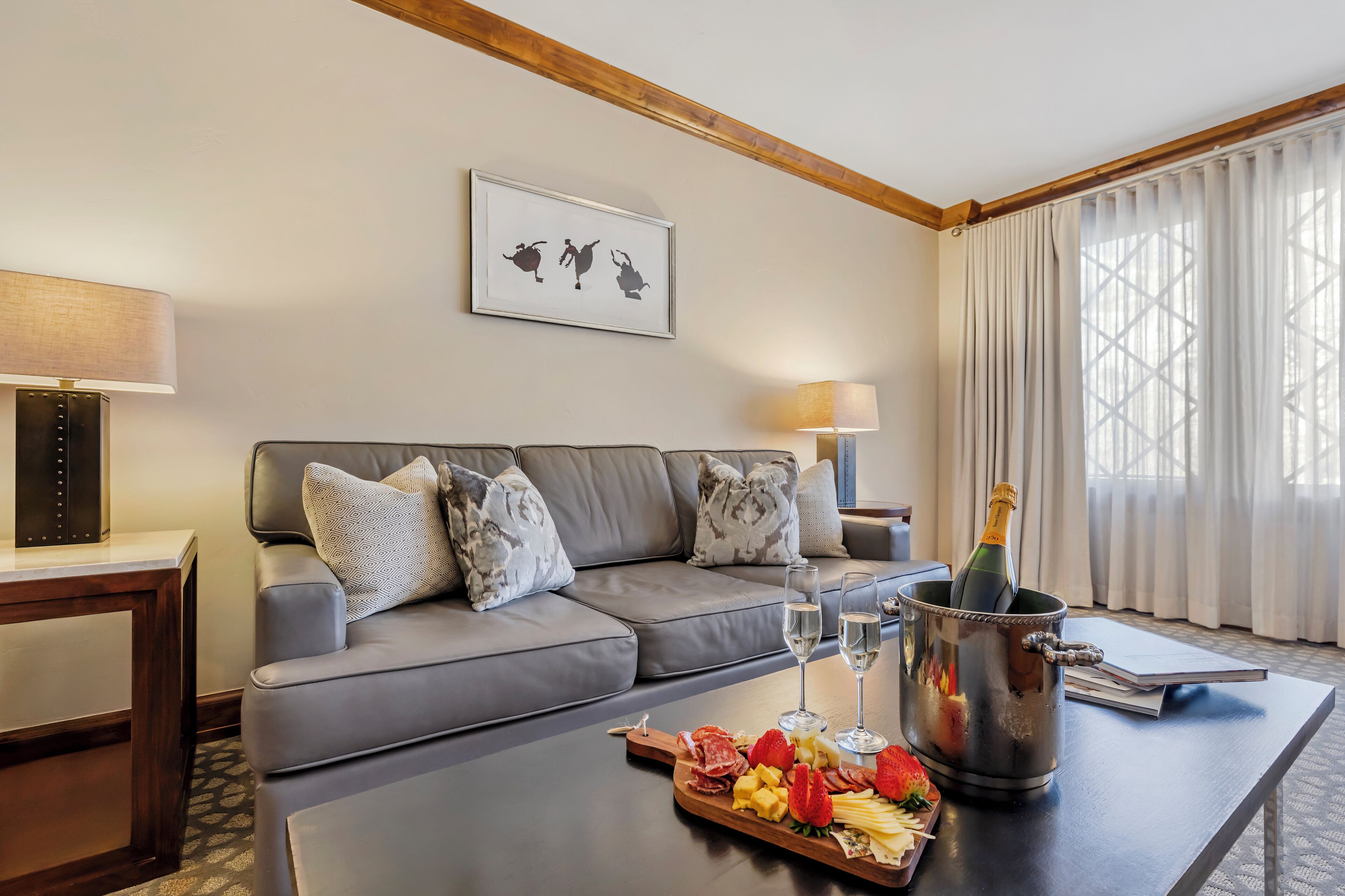 A modern living room with a grey sofa, two side tables with lamps, a coffee table with a bottle of champagne, glasses, and a fruit platter.