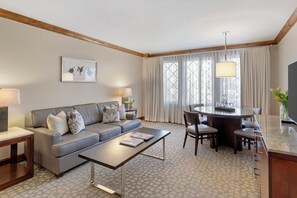 A modern hotel room with a grey sofa, a coffee table, a dining area with a round table and chairs, and a large window with curtains.